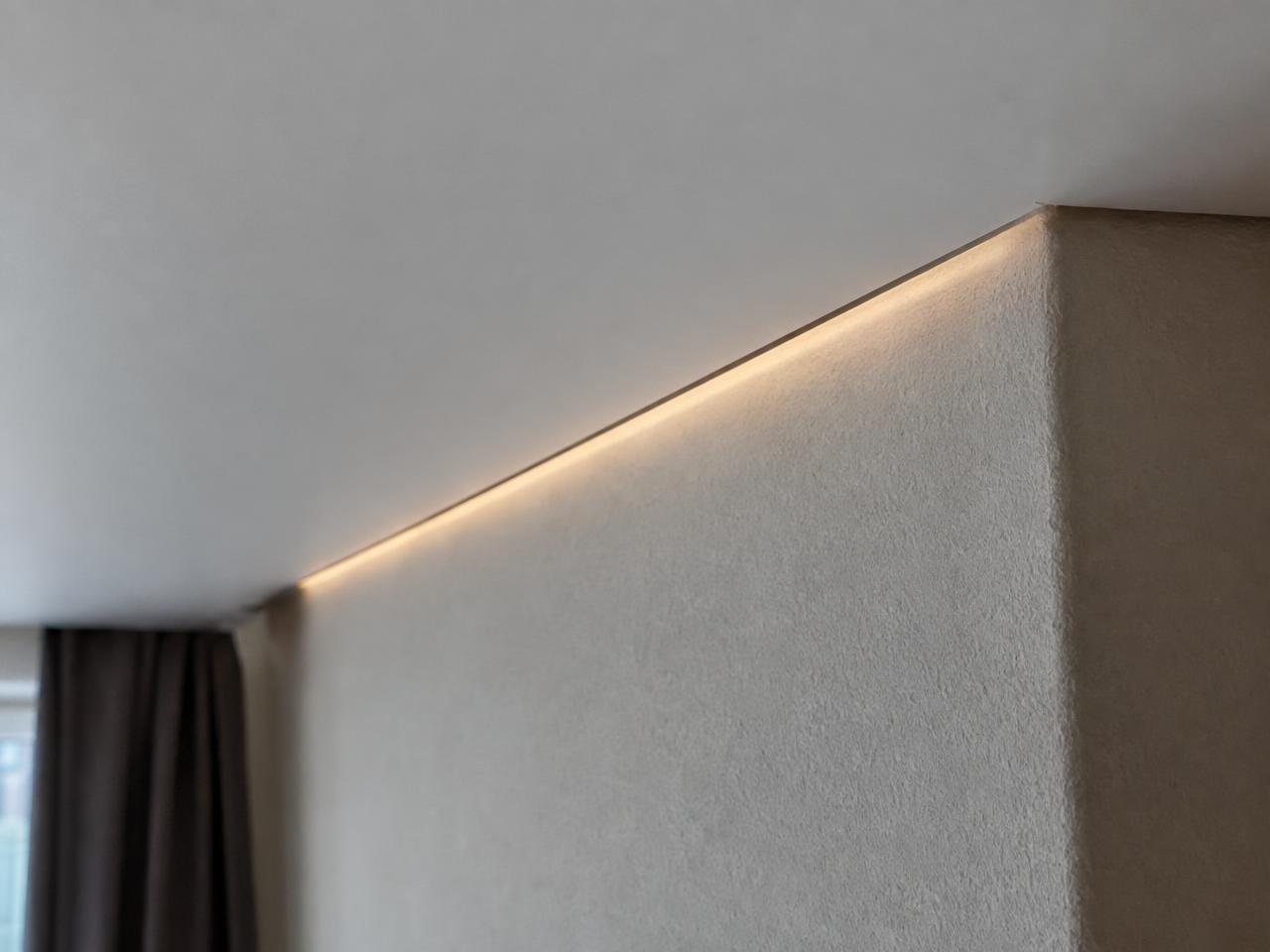 Close-up architectural detail photograph of the corner where a matte white stretch ceiling meets a painted wall through a 10 mm shadow gap, a soft line of warm light inside the gap, precise and clean, shallow depth of field. Documentary photograph, real location in Russia, natural imperfect details, true-to-life colours, soft film grain, no text, no logos, no watermark, no CGI look, no glossy plastic skin, no lens flare, no neon, no holograms.