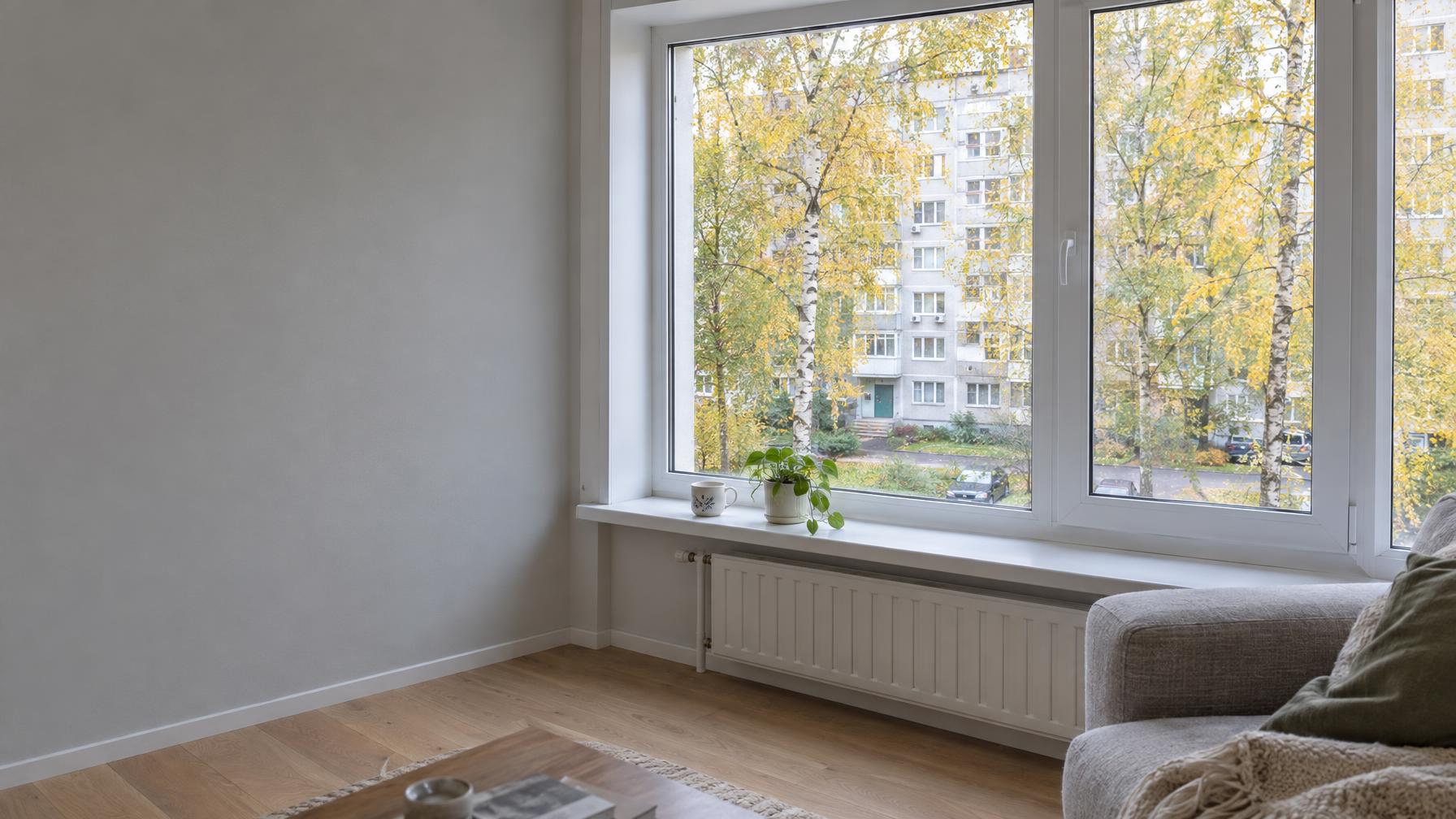 Wide real-estate photograph of a bright living room in a renovated Moscow panel-block apartment: a large new white PVC tilt-and-turn window with thin frame, view onto a courtyard with yellow birches in early autumn, soft morning daylight, clean windowsill with a ceramic mug and a small plant, light oak floor. Shot on 24mm at eye level, calm and airy. The window is on the right two thirds of the frame; the left third is a calm light-grey wall — empty space for a headline. Documentary photograph, real location in Russia, natural imperfect details, true-to-life colours, soft film grain, no text, no logos, no watermark, no CGI look, no glossy plastic skin, no lens flare, no neon, no holograms.
