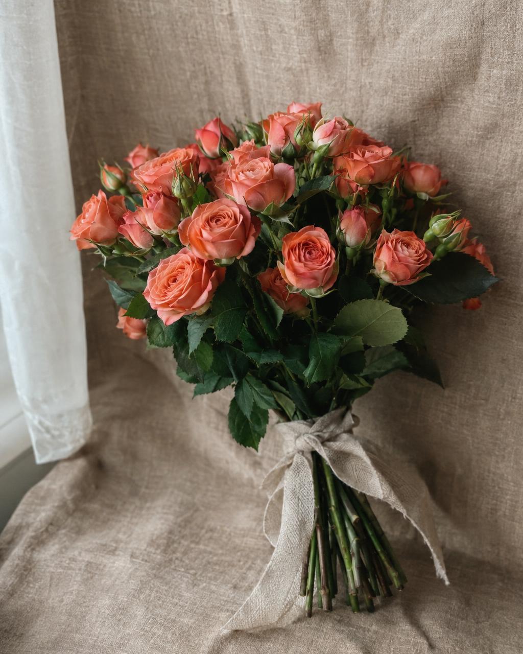 Product photograph of a compact bouquet of coral spray roses tied with a linen ribbon, on a warm beige linen backdrop, soft window light, real leaves. Documentary photograph, real location in Russia, natural imperfect details, true-to-life colours, soft film grain, no text, no logos, no watermark, no CGI look, no glossy plastic skin, no lens flare, no neon, no holograms.